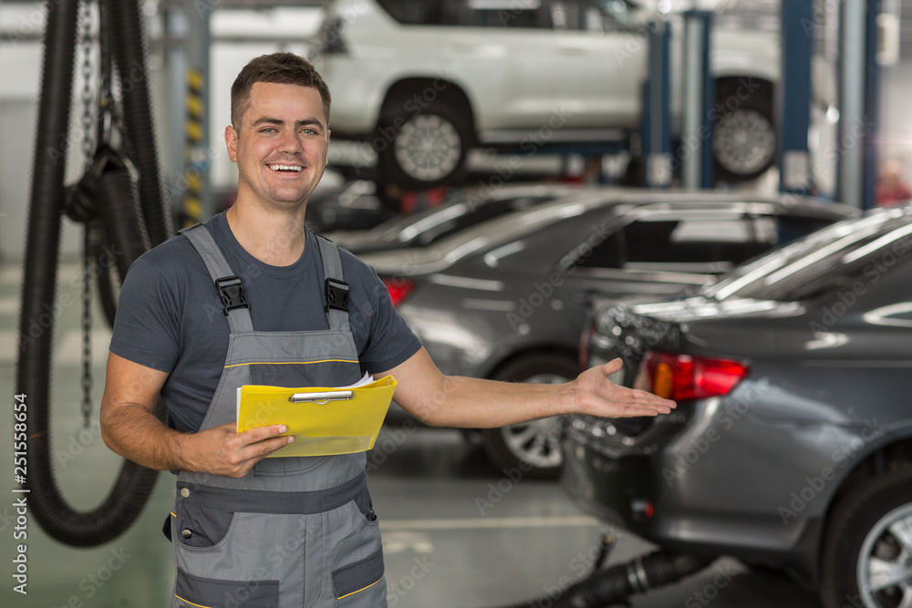 Portrait of positive repairman in uniform showing with hand auto service equipped with modern technology. Smiling mechanic keeping folder, gesturing with hand on cars and looking at camera. Portrait of positive repairman in uniform showing with hand auto service equipped with modern technology. Smiling mechanic keeping folder, gesturing with hand on cars and looking at camera.