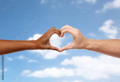 charity, love and diversity concept - close up of female and male hands of different skin color making heart shape over blue sky and clouds background