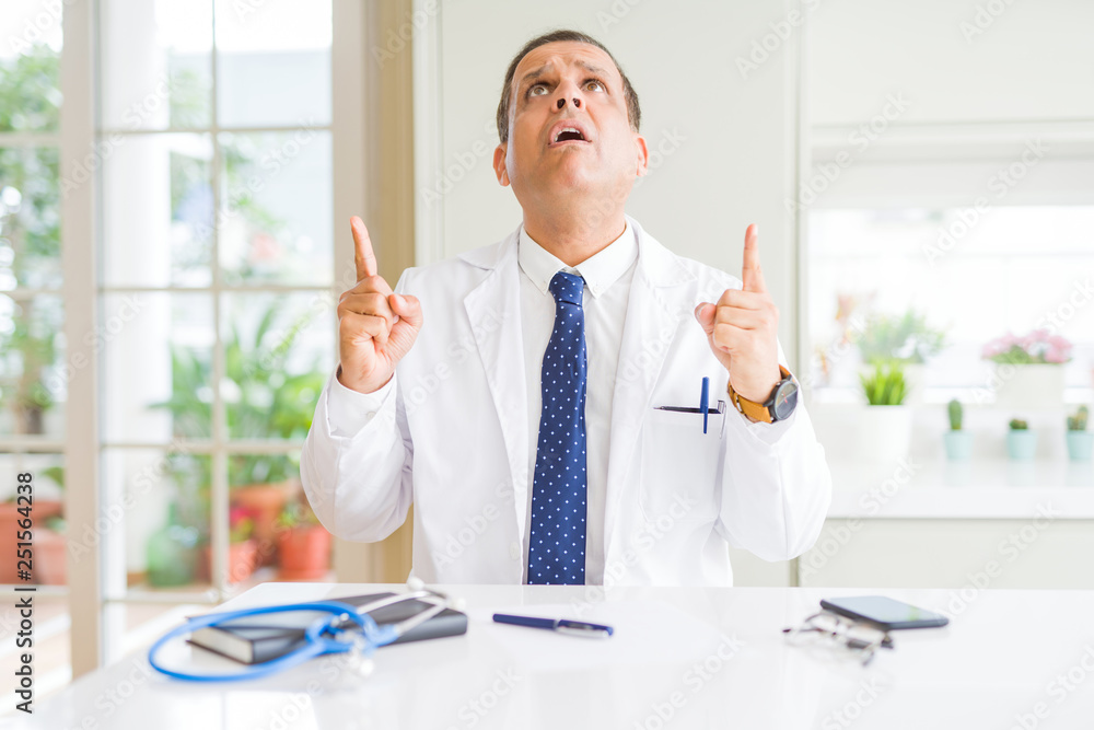 Middle age doctor man wearing medical coat at the clinic amazed and surprised looking up and pointing with fingers and raised arms. Middle age doctor man wearing medical coat at the clinic amazed and surprised looking up and pointing with fingers and raised arms.