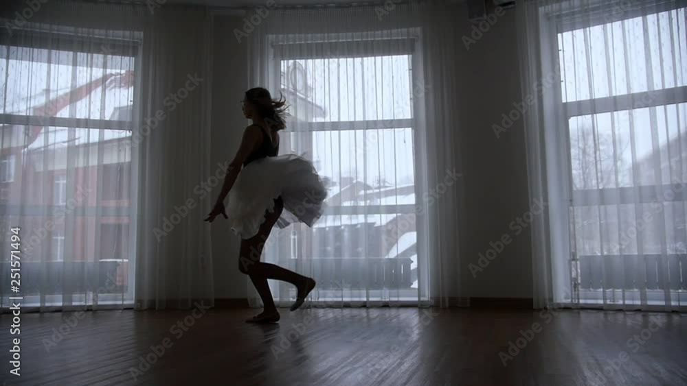 A studio in twilight. A silhouette of young woman ballerina runs up and jumps performing a split in the air. A studio in twilight. A silhouette of young woman ballerina runs up and jumps performing a split in the air.