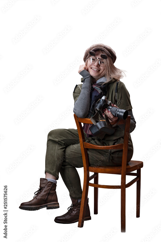 Young woman in a green jumpsuit, a cap and glasses sits on a chair with a retro camera isolated on a white background.