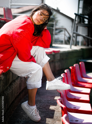 Young normal beautiful tennager girl model in the seats of a baseball stadium wearing red bomber jacket, hipster rounded glasses, white pants and sneakers