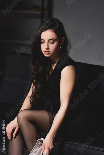 attractive brunette woman in elegant dress sitting on couch on dark background © LIGHTFIELD STUDIOS