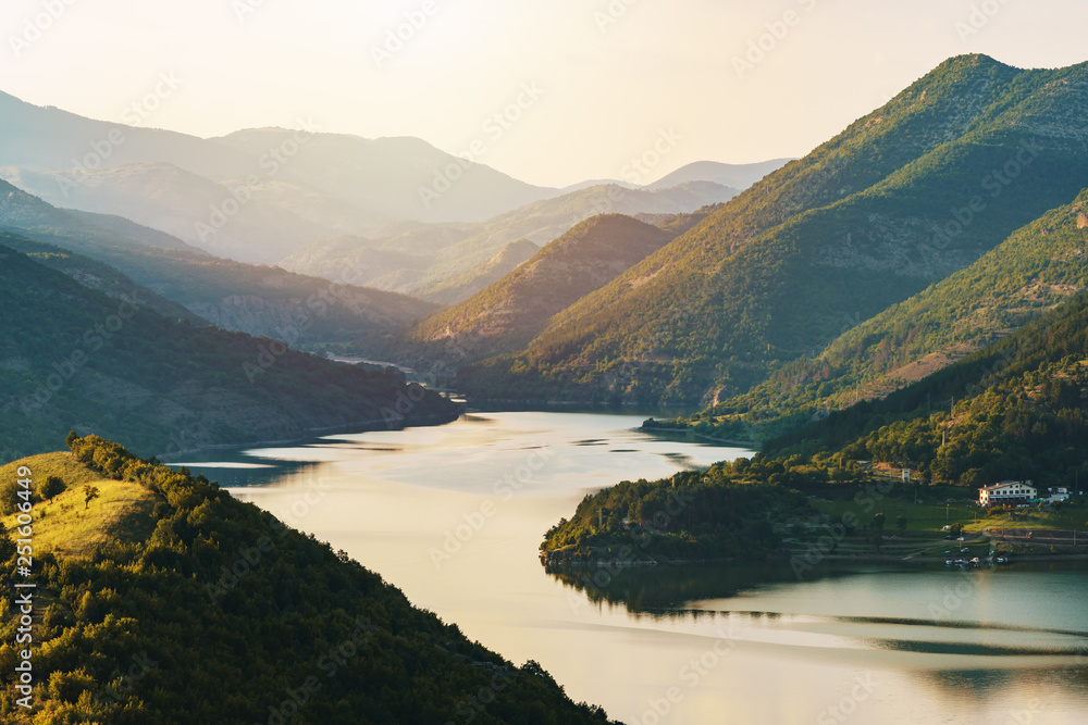 Picturesque bright panorama of the mountains in the summer in the light of sunset. Mountain gorge with a lake and a pine forest along the shore. Orange light in the mountains. Relax tourist