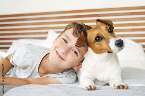 Happy boy playing with his dog, Jack Russell Terrier, waking up early in the morning, in a bed in white bedding. Smiling child and his pet basking in bed. Resort vacation at the hotel