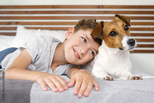 Happy boy playing with his dog, Jack Russell Terrier, waking up early in the morning, in a bed in white bedding. Smiling child and his pet basking in bed. Resort vacation at the hotel