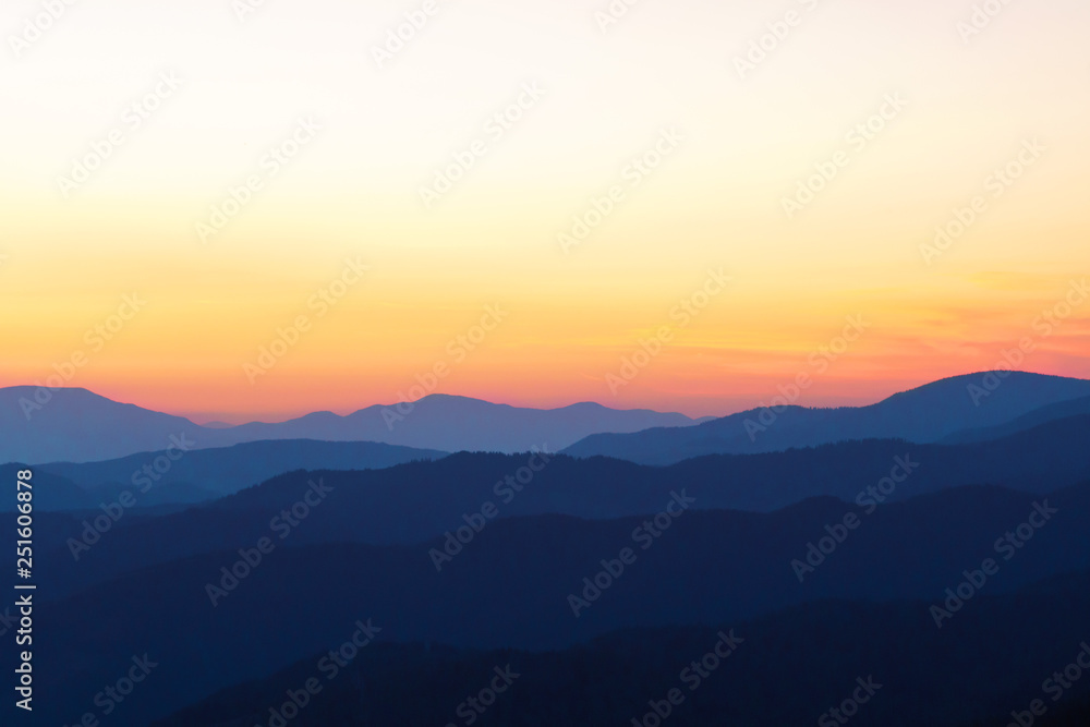 Panorama of the peaks of beautiful mountains covered with trees at sunset on a sunny day in summer. Orange glow on the sky after full sunset. Dark silhouettes of mountains. Solar reflection