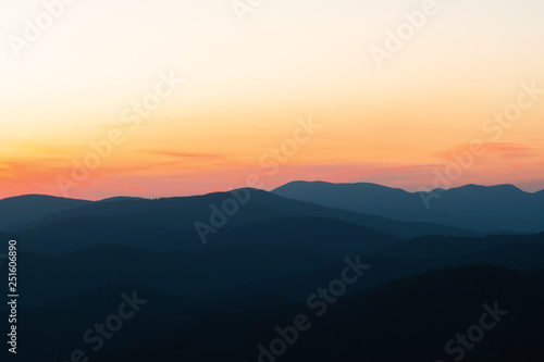 Panorama of the peaks of beautiful mountains covered with trees at sunset on a sunny day in summer. Orange glow on the sky after full sunset. Dark silhouettes of mountains. Solar reflection