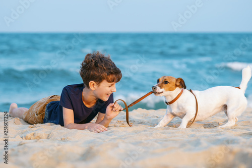 Happy boy hugging his dog breed Jack Russell terrier at the seashore against a blue sky close up at sunset. Best friends rest and have fun on vacation, play in the sand against the sea summer