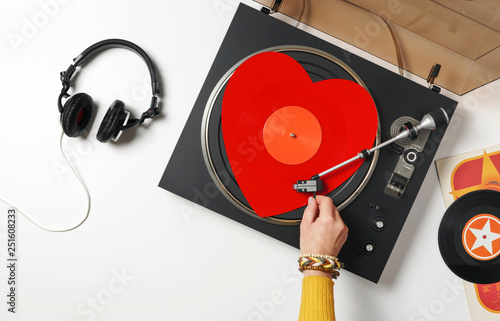 A red heart vinyl record turntable on a white table with plates. Included gramophone, black headphones. The hand of a girl DJ with bright accessory puts a stylus on a vinyl record. Valentine's Day photo