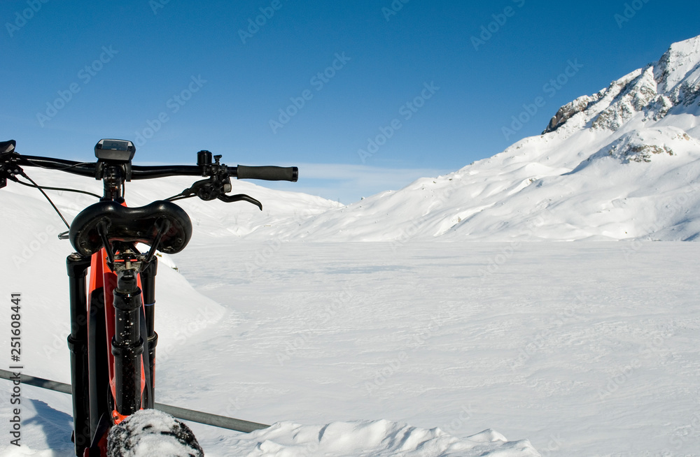 orange electric bicycle, e-bike, ebike, snowy mountains of Formazza Valley and frozen lake of Toggia, winter, sport, adventure, travel, Alps, Piedmont, italy