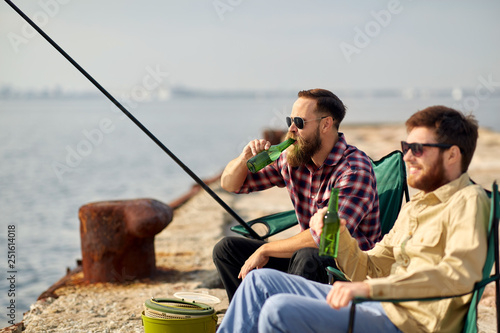 leisure and people concept - happy friends fishing and drinking beer on pier