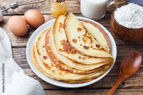 Thin pancakes on a plate. Wooden background. Ingredients to cooking eggs, milk, flour.