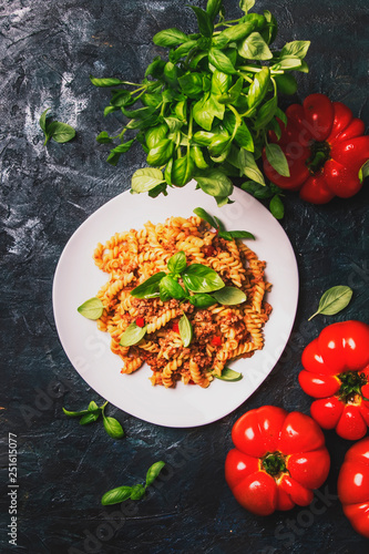Tasty pasta with minced meat and tomato sauce with green basil on white plate, dark background kitchen table, top view