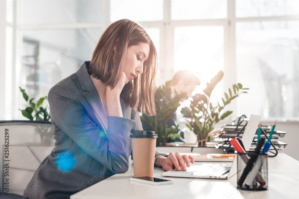selective focus of pensive businesswoman using laptop near paper cup with coworker on background selective focus of pensive businesswoman using laptop near paper cup with coworker on background
