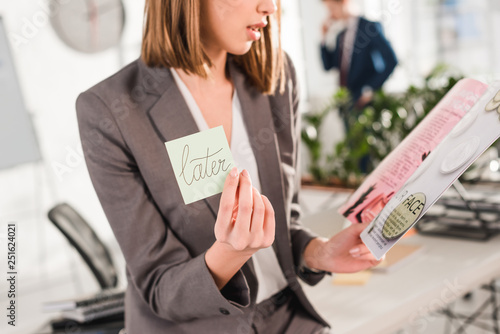 cropped view of businesswoman holding sticky note with later lettering and magazine in hands with coworker on background, procrastination concept