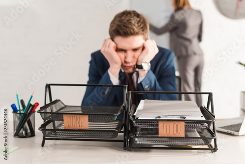 selective focus of document trays with lettering near tired man and female coworker on background, procrastination concept photo