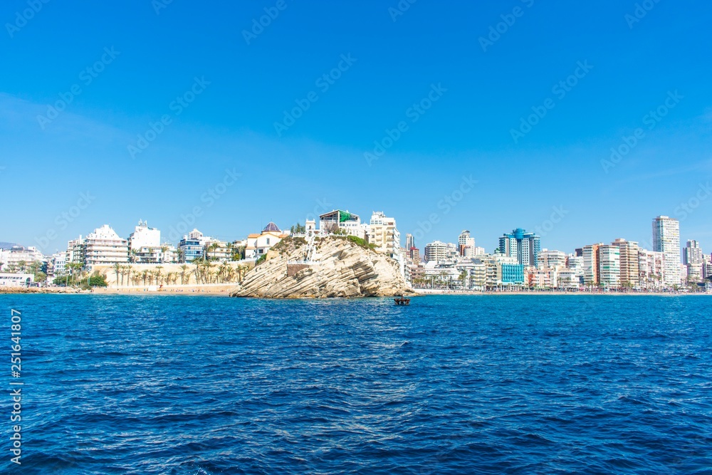 A beautiful rock protruding from golden sandy beaches on the coastline of Benidorm, Spain, with touristy buildings and skyscrapers in the background