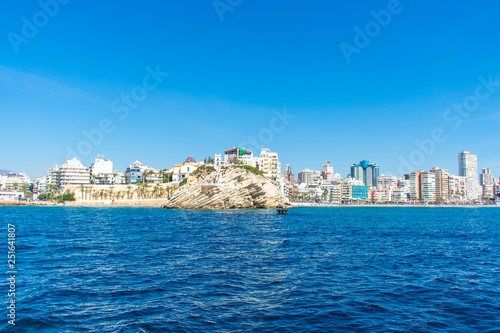 A beautiful rock protruding from golden sandy beaches on the coastline of Benidorm, Spain, with touristy buildings and skyscrapers in the background © JC