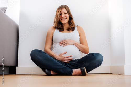 joyful pregnant woman in white top sitting on the ground holding her belly