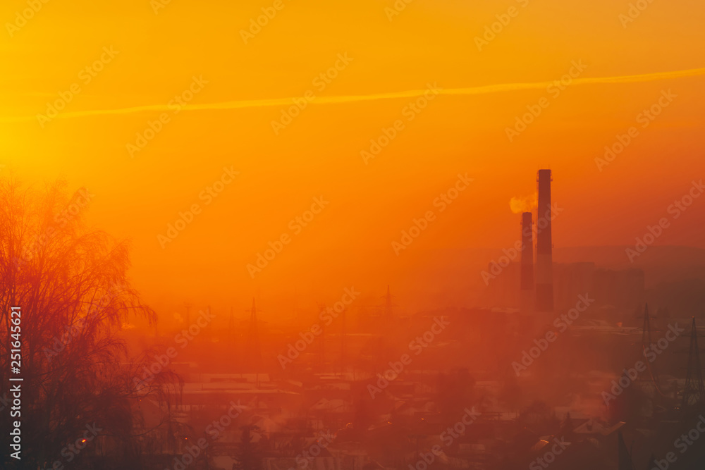 Smog among silhouettes of buildings on sunrise. Smokestack in dawn sky. Environmental pollution on sunset. Harmful fumes from stack above city. Mist urban background with warm orange yellow sky.