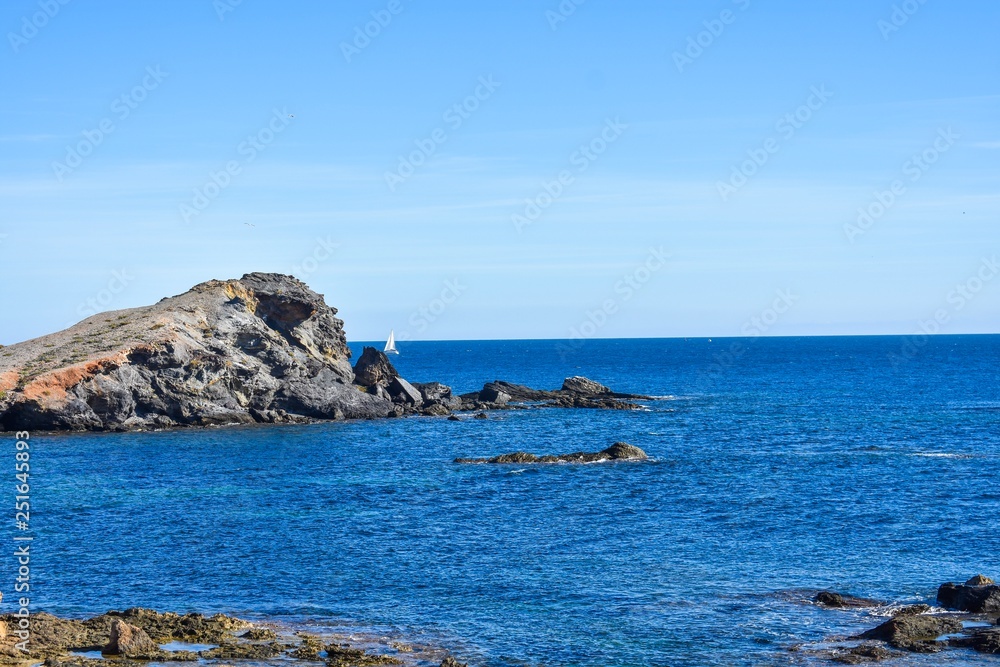 The rocky shore of the south of Spain looking out over a small sailboat on the blue water of the Mediterranean
