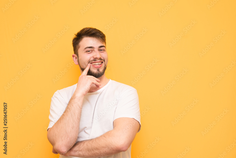 Happy man with a beard wears a white T-shirt, stands on a yellow background, looks into the camera and smiles.Smiling man holds his finger around the mouth, isolated on a yellow background. Copyspace Happy man with a beard wears a white T-shirt, stands on a yellow background, looks into the camera and smiles.Smiling man holds his finger around the mouth, isolated on a yellow background. Copyspace