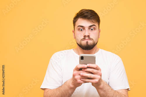 Funny man dressed in a white T-shirt use smartphone and sad looking up at the screen.Man with a beard focused on looking at the screen and experiencing emotions,isolated on a yellow background © bodnarphoto