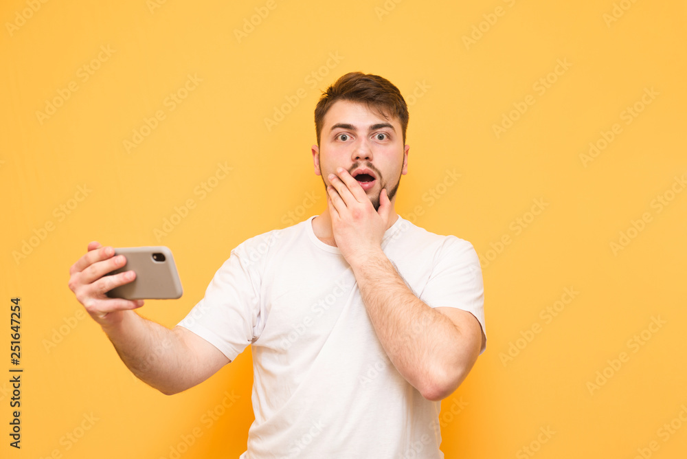 Shocked man with a beard standing on a yellow background with a smartphone , looks at the camera with astonishment. Amazed gamer with a smartphone in his hands is isolated on a yellow background.