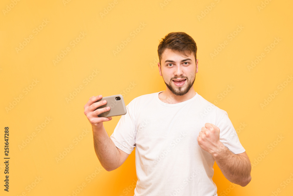 Happy man with a smartphone in his hand, wearing a white T-shirt, looking into the camera and rejoicing. Joyful man with a beard uses a smartphone, isolated on a yellow background. Copyspace Happy man with a smartphone in his hand, wearing a white T-shirt, looking into the camera and rejoicing. Joyful man with a beard uses a smartphone, isolated on a yellow background. Copyspace