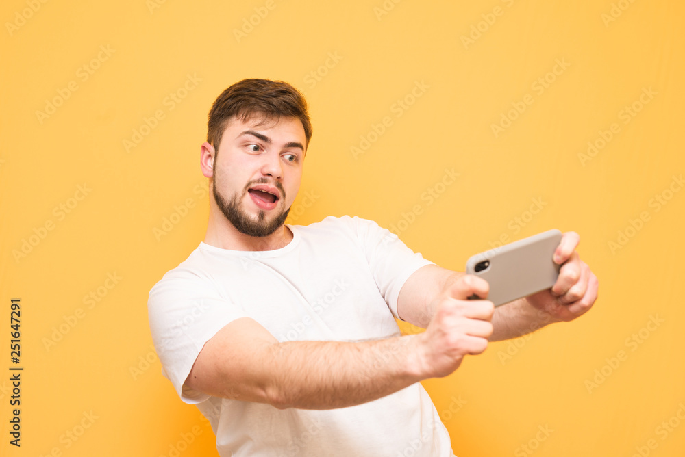Happy adult man with a beard expressively playing video games on a smartphone on a yellow background. Joyful gamer in a white T-shirt plays mobile games on a colored background. Copyspace Happy adult man with a beard expressively playing video games on a smartphone on a yellow background. Joyful gamer in a white T-shirt plays mobile games on a colored background. Copyspace