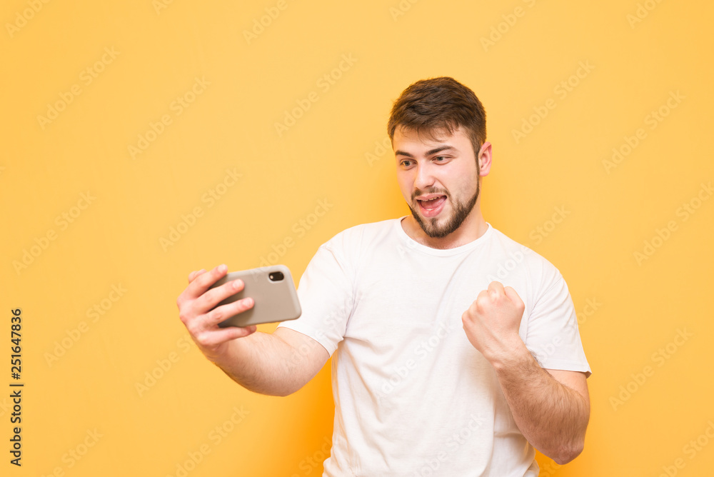 Adult man with a beard playing mobile games on a smartphone, looking at the screen and rejoicing, wearing a T-shirt, standing on a yellow background. Mobile gaming. Adult man with a beard playing mobile games on a smartphone, looking at the screen and rejoicing, wearing a T-shirt, standing on a yellow background. Mobile gaming.