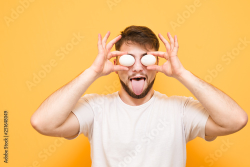 Funny man holds eggs instead of his eyes and shows a tongue on a yellow background. Positive Man with Beard keeps chicken eggs at eye level.