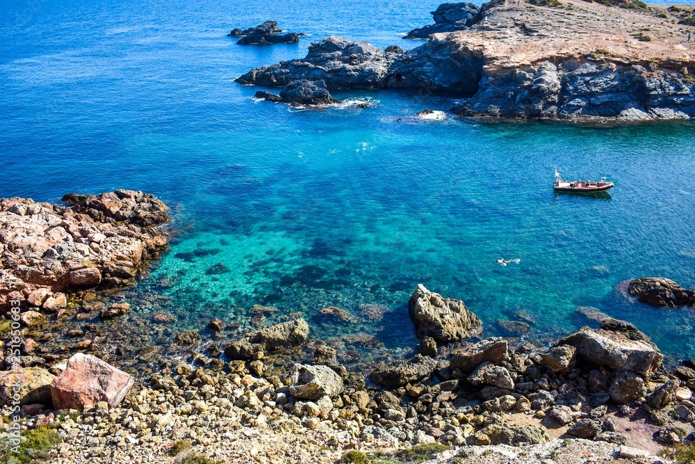 Crystal clear water envelop dark grey rocks that just rise above the surface, with a small boat and some people snorkelling.