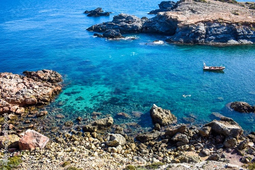 Crystal clear water envelop dark grey rocks that just rise above the surface, with a small boat and some people snorkelling.