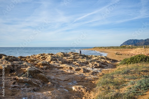 The dry semi-desert of southern Spain meets the blue water of the mediterranean with a rocky foreground and a sandy beach in the background, along the coast of southern Spain