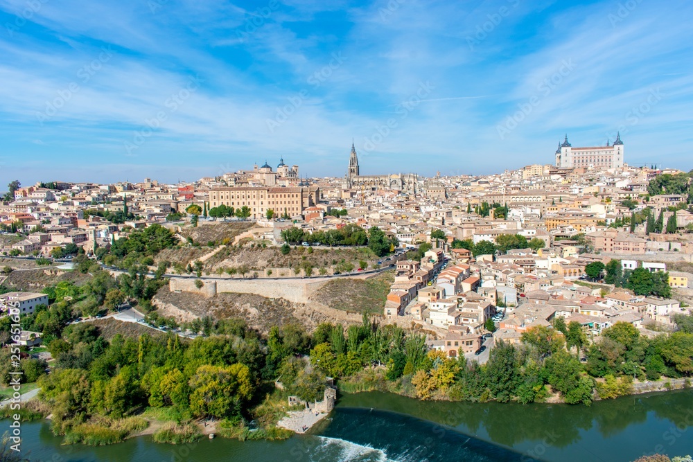 A panoramic view of the old city of Toledo, with it’s defensive walls, the tower of the cathedral and the old castle on top a hill inside the city walls, surrounded by a river that serves as a moat.