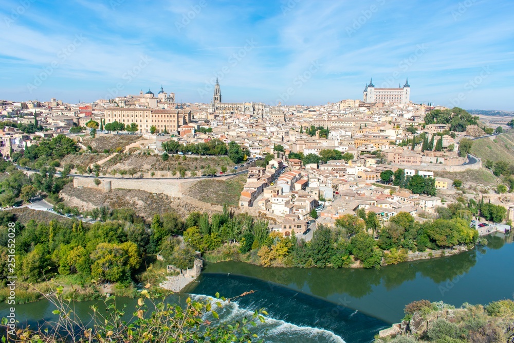 A panoramic view of the old city of Toledo, with it’s defensive walls, the tower of the cathedral and the old castle on top a hill inside the city walls, surrounded by a river that serves as a moat.