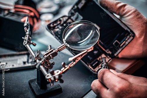 Repairman uses magnifier and tweezers to repair damaged smartphone. Close-up photo of a disassembled smartphone.