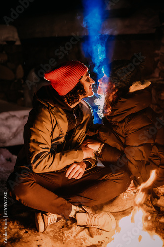 Stunning guy and the girl are sitting near the fire and exhale blue smoke. Romantic night