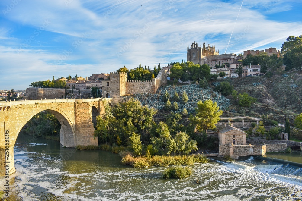 A view of an ancient bridge over the river moat of Toledo, Spain, with it’s old fortified gatehouse and houses on the far side creeping up the hill to where the ancient monastery church is and lush gr