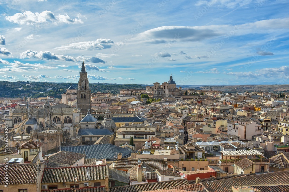A tiled rooftop view of Toledo, Spain with the towers of the cathedral and monastery peaking out from amongst the closely spaced buildings. 