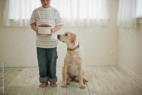 Young boy holding bowl of dog food as his dog looks up at it. photo