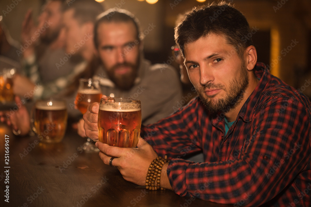Client of beer pub sitting and resting. Handsome bearded man in checked shirt looking at camera and holding glass of tasty beer. People in pub communicating and socializing. Client of beer pub sitting and resting. Handsome bearded man in checked shirt looking at camera and holding glass of tasty beer. People in pub communicating and socializing.