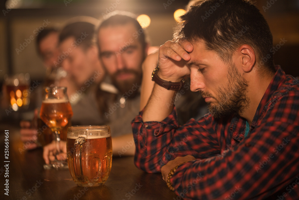 Sad and depressed facial expression of bearded man looking down. Male client sitting in beer pub and holding hand on head. People resting in bar and communicating on background. Sad and depressed facial expression of bearded man looking down. Male client sitting in beer pub and holding hand on head. People resting in bar and communicating on background.