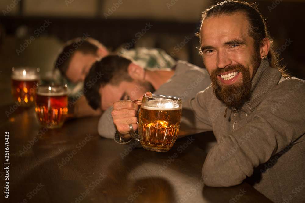 Side view of positive and happy bearded; handsome man holding glass of beer and drinking. Man enjoying and tasting delicious beer looking and smiling at camera. People resting in pub. Side view of positive and happy bearded; handsome man holding glass of beer and drinking. Man enjoying and tasting delicious beer looking and smiling at camera. People resting in pub.