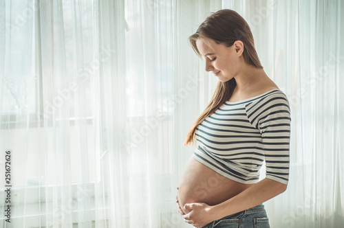 Happy Attractive pregnant woman standing near the window and holding her belly