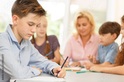 Boy with serious facial expression, sitting at desk and drawing pencil on paper.