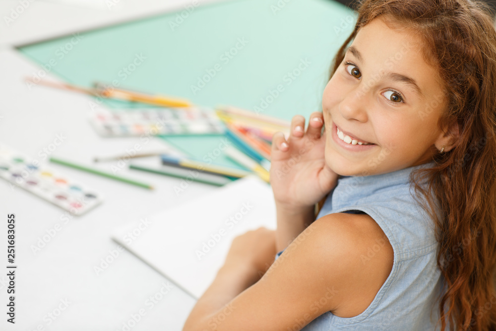 View over shoulder of adorable little red haired girl sitting conceived at school desk at classroom. Female child, wearing in blue shirt smiling, looking at camera, when drawing.