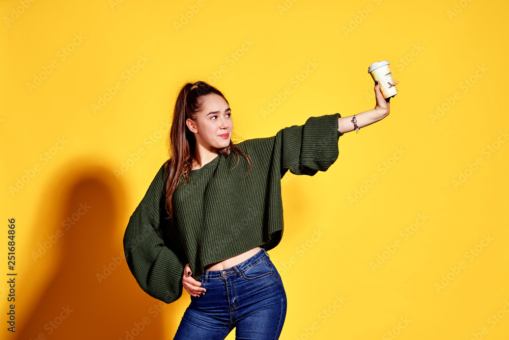 Image of pretty young woman smiling and holding takeaway coffee in paper cup isolated over yellow background Image of pretty young woman smiling and holding takeaway coffee in paper cup isolated over yellow background